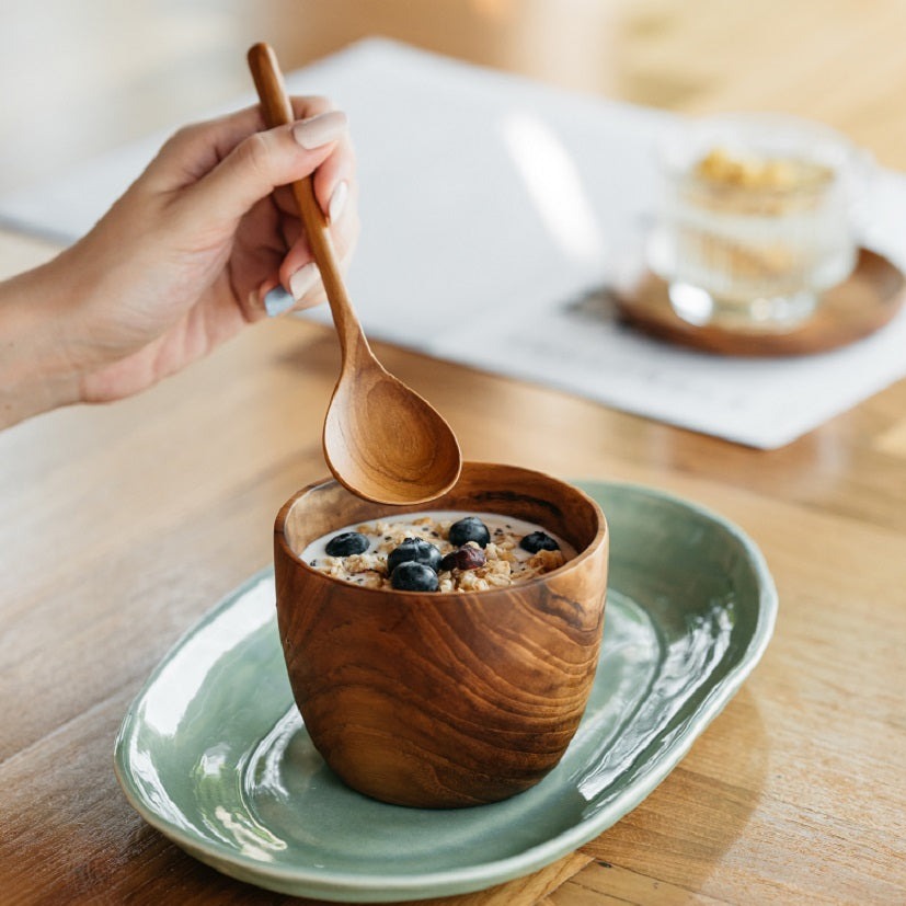 A person is holding a wooden spoon above a bowl of oatmeal topped with blueberries. The bowl is placed on a green plate on a wooden table.