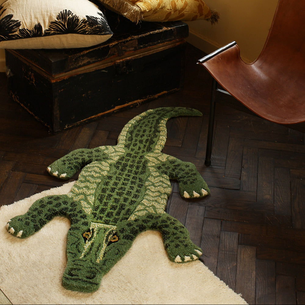 Green alligator-shaped rug on a wooden floor with pillows and a chair in the background.