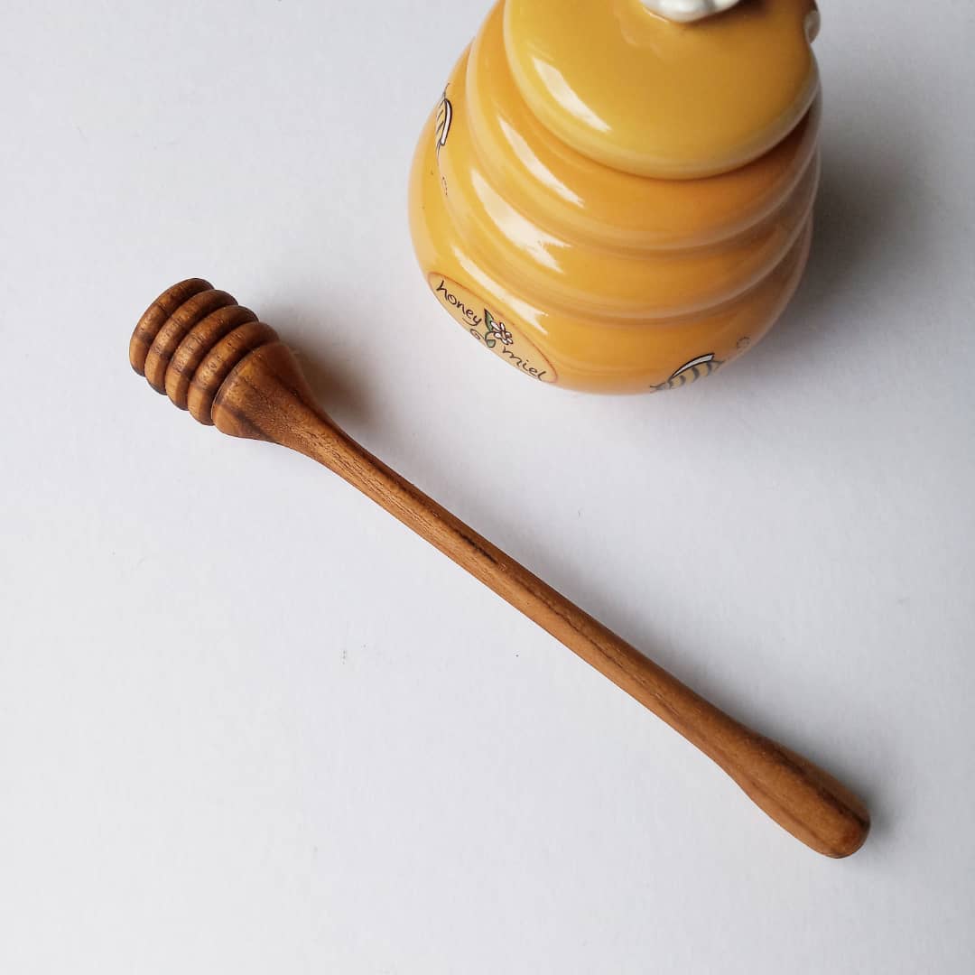 A handcrafted Teak Wood honey dipper lying on a white surface, next to a yellow honey jar with a floral pattern.