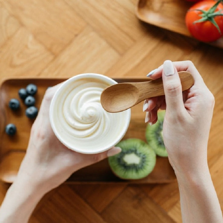 A person holding a teak wood ice cream spoon above a bowl of ice cream, with fruits and a wooden board in the background.