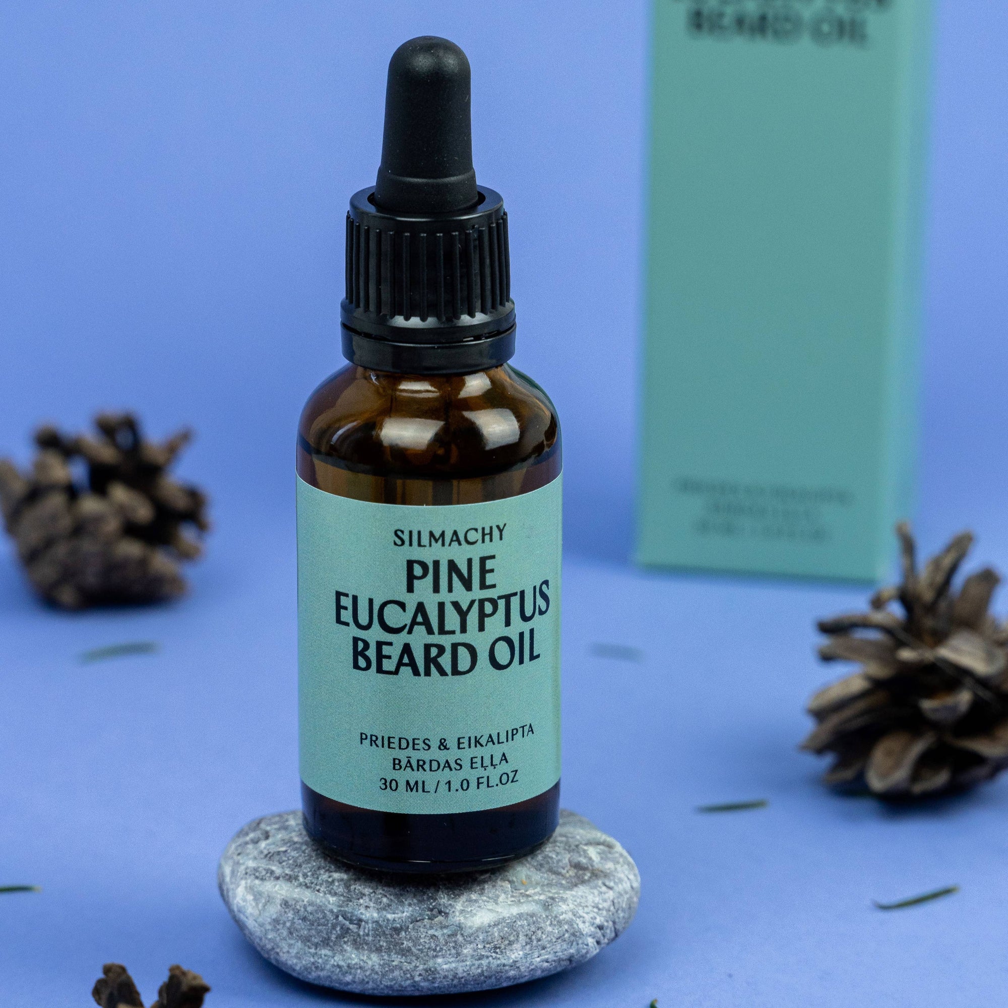 Bottle of Silmachy Pine Eucalyptus Beard Oil on a blue background with pine cones.