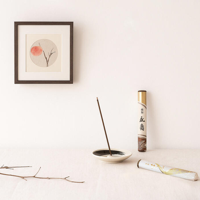 Incense stick and holder with a roll of incense on a light surface, framed artwork in the background.