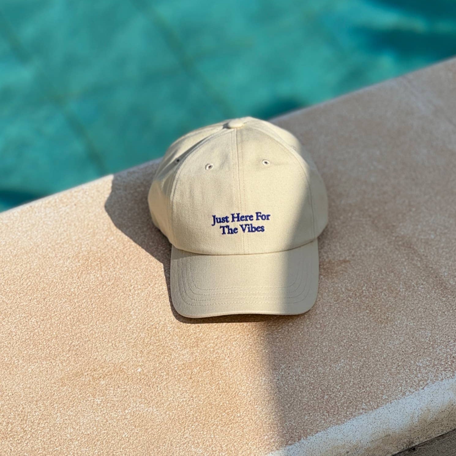Beige cap with 'Just Here For The Vibes' text on a pool ledge