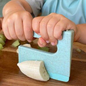 Child using a blue banana slicer on a wooden table with green bananas.