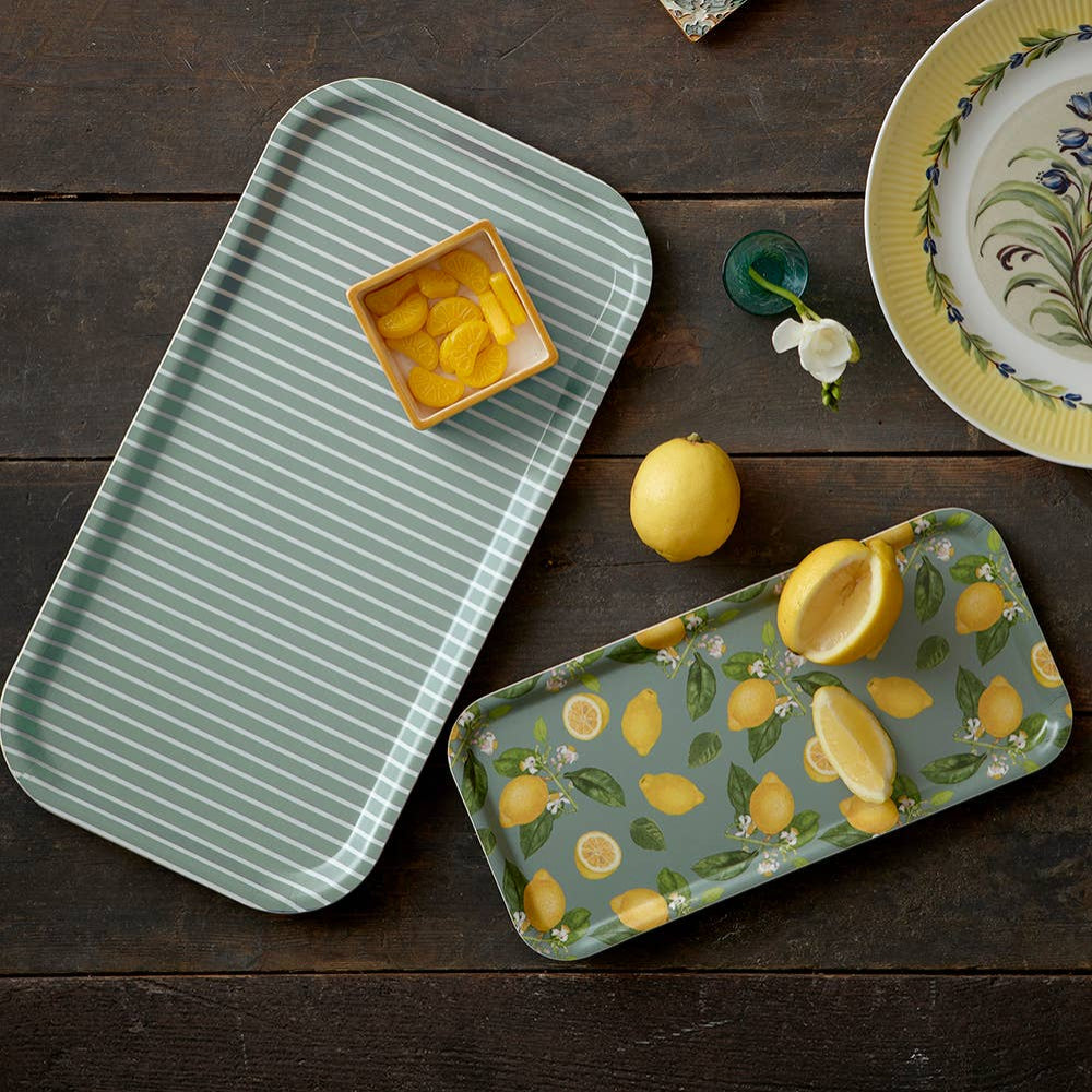 Two trays on a wooden surface, one with green stripes and the other with lemon design, along with lemons and a bowl.
