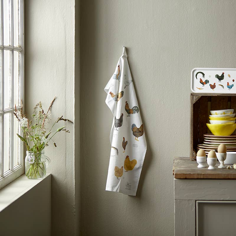 Kitchen towel with chicken design hanging on a wall next to a window with flowers and kitchenware.