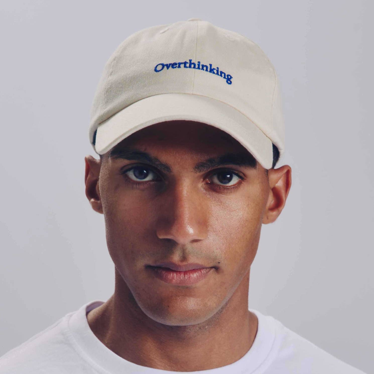 Person wearing a beige cap with 'Overthinking' text on a plain background