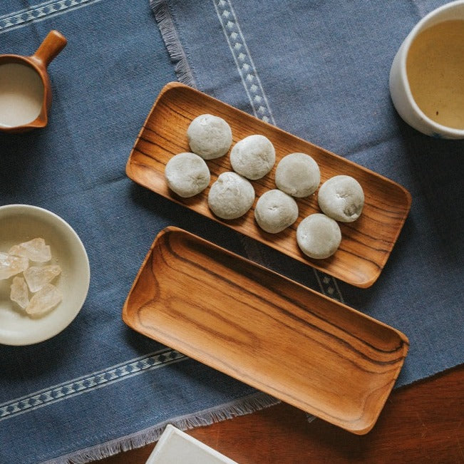 A rectangular teak wood plate placed on a table with a blue textured cloth, displaying various food items like rolls and sweets.