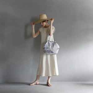 Woman in a white dress and straw hat holding a patterned bag against a gray background