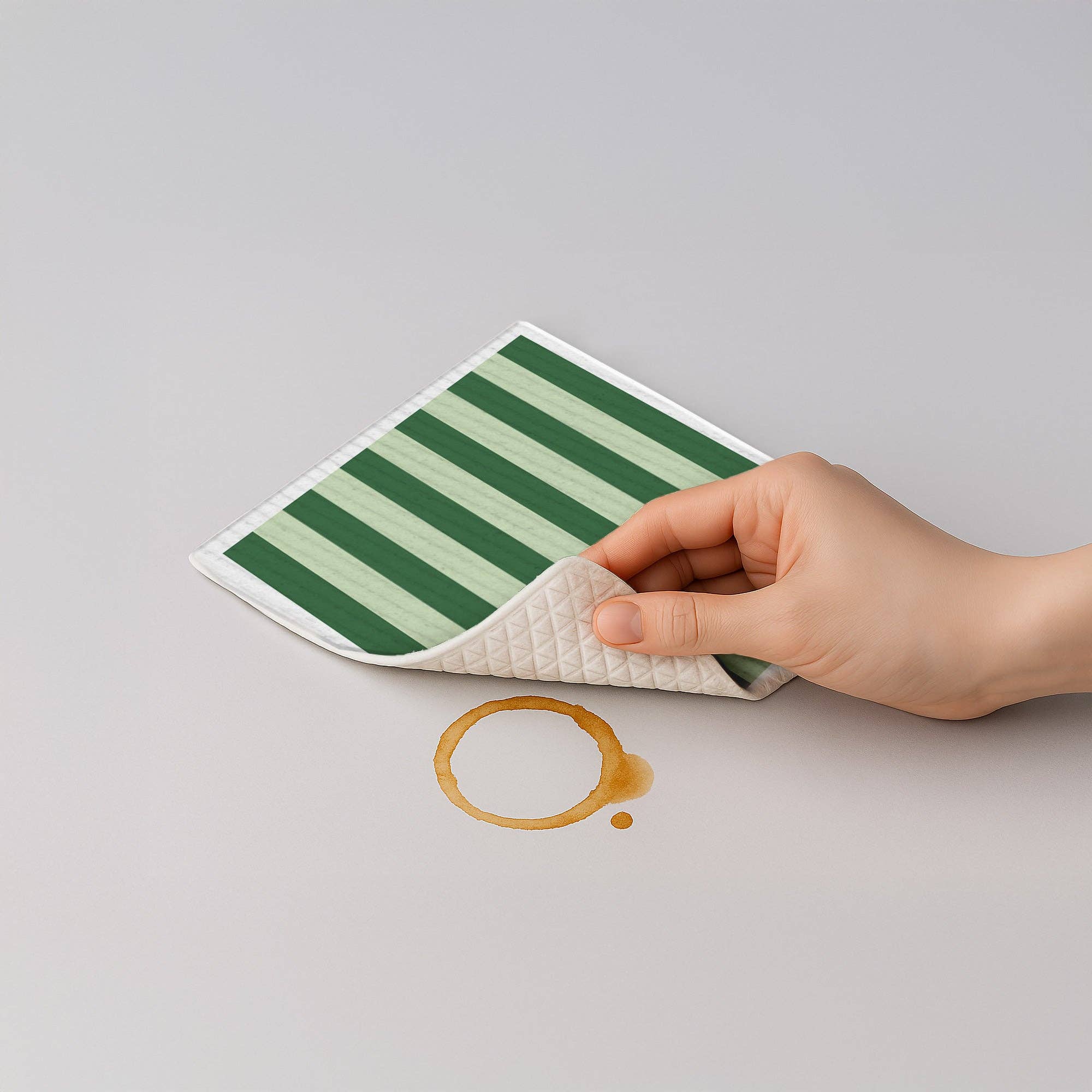 Hand holding a green and white striped mat over a coffee spill on a gray surface