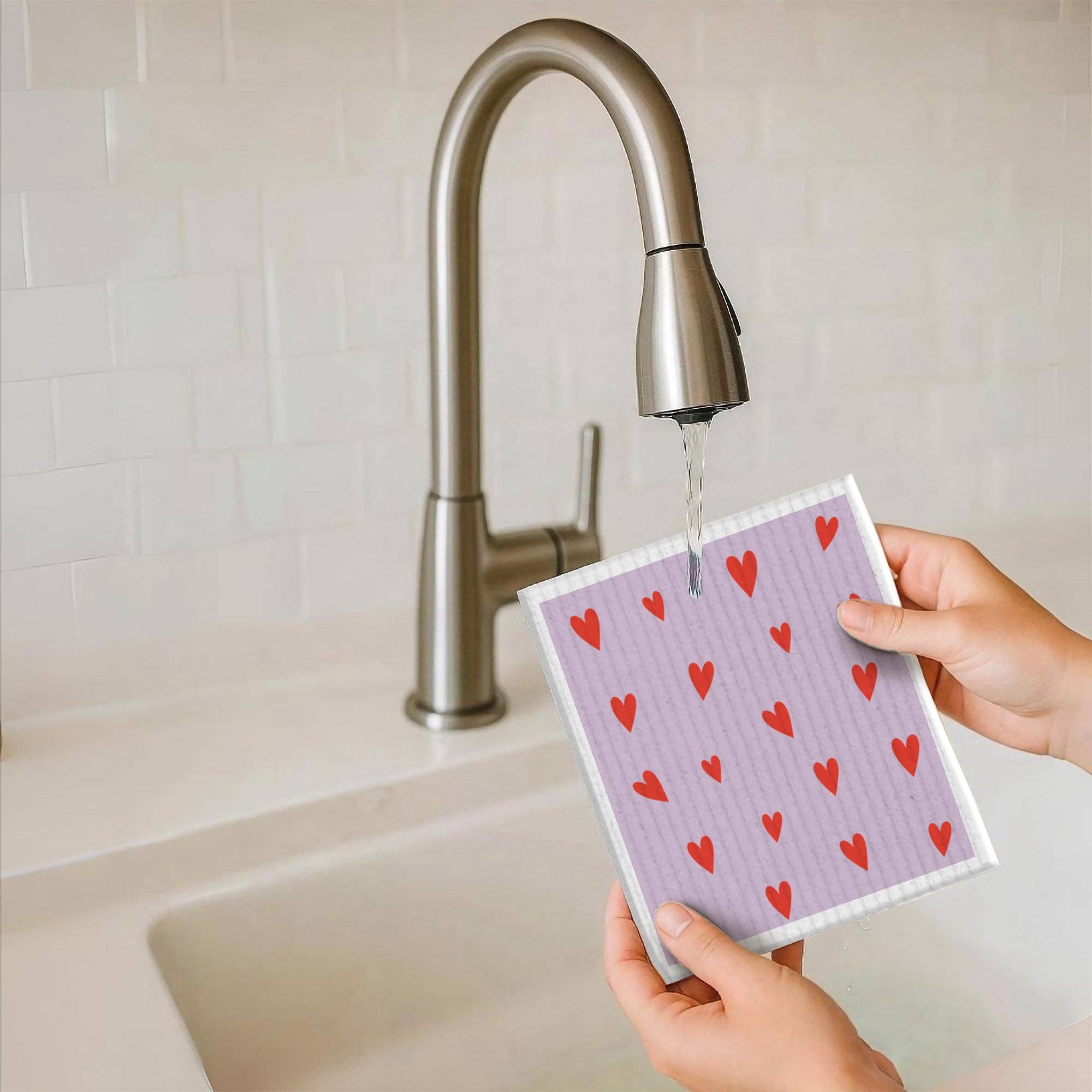 Person holding a dishcloth with red heart pattern under running water in a kitchen.