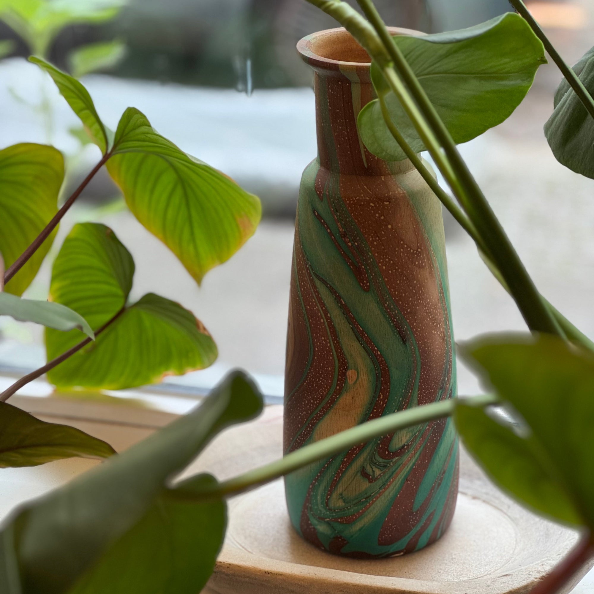 Decorative vase with marbled pattern on a wooden stand surrounded by green leaves.