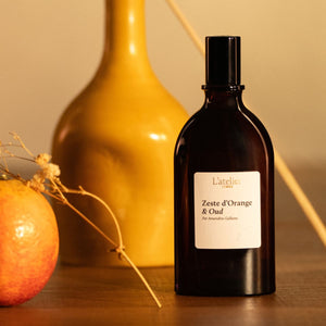 Black bottle of 'Zeste d'Orange & Oud' by L'Atelier on a wooden surface with an orange and vase in the background.