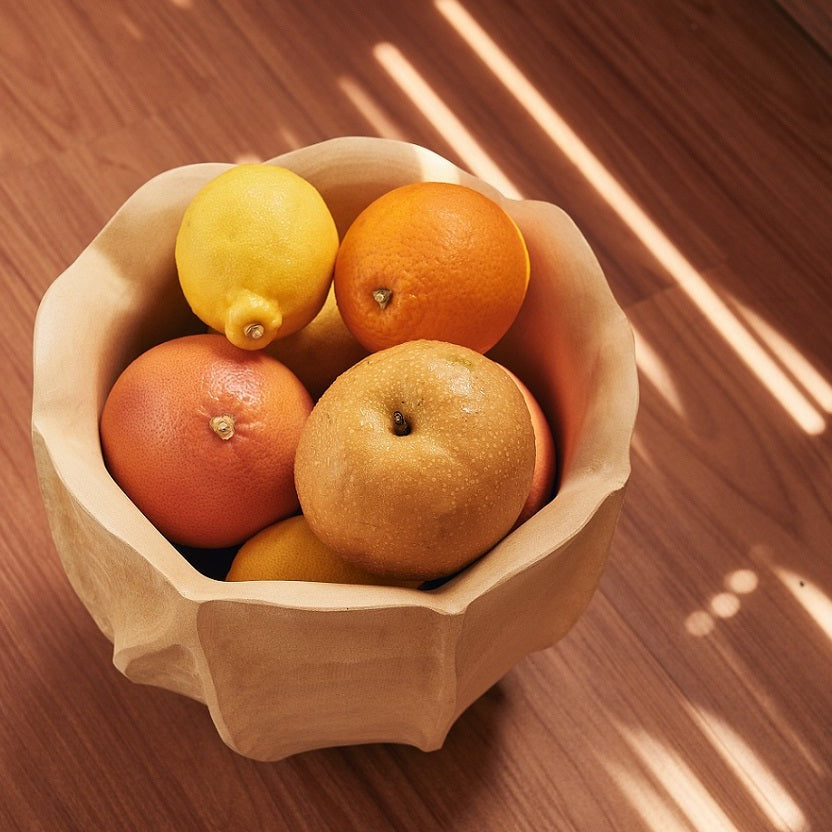A handcrafted mango wood fruit bowl on a table, filled with various fruits like oranges and apples.