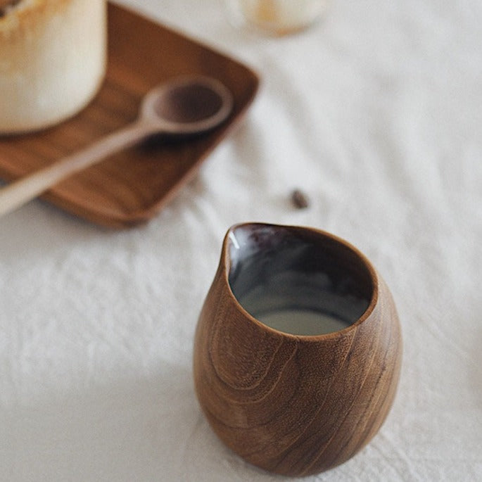 A small teak wood milk pitcher with a dark brown color and a smooth finish, placed on a white surface, with a wooden board and a spoon in the background.