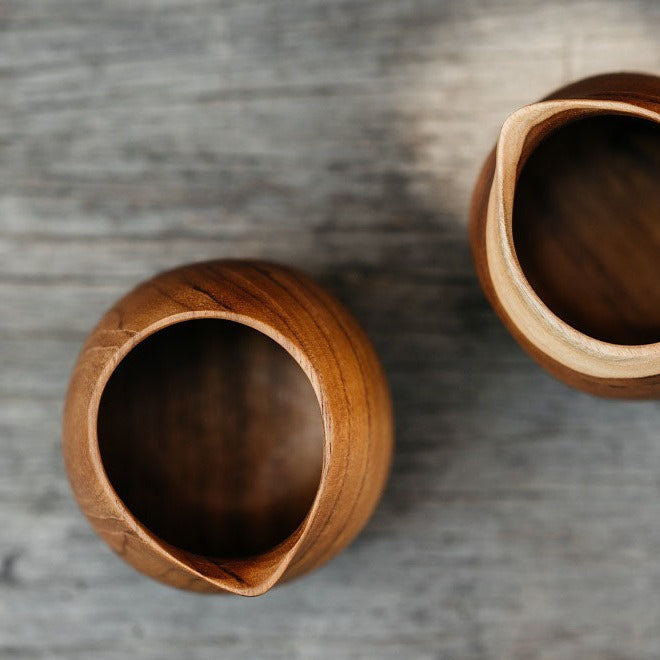 A small teak wood milk pitcher with a dark brown color and a smooth finish, placed on a white surface, with a wooden board and a spoon in the background.