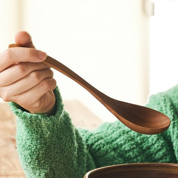 A person holding a brown Teak Wood Ramen Spoon over a bowl, with a soft-focus background.