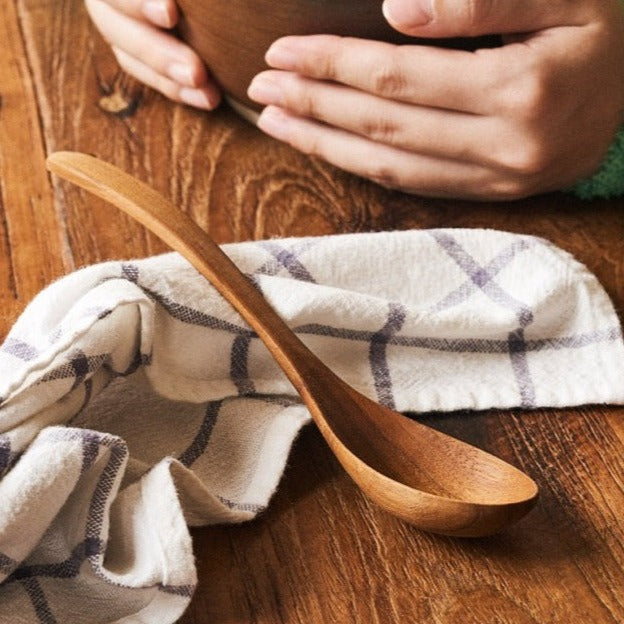 A person holding a brown Teak Wood Ramen Spoon over a bowl, with a soft-focus background.