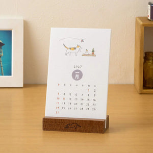 A 2027 monthly desk calendar with a minimalist cat illustration stands on a wooden block base on a light wood tabletop. A framed picture and a small wooden shelf with a bottle are blurred in the background against a beige wall.