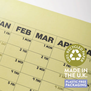 A close-up of a pale yellow monthly calendar page with black printed dates and headers, plus a circular green '100% Recycled Paper' badge and a blue 'Plastic Free Packaging' label.