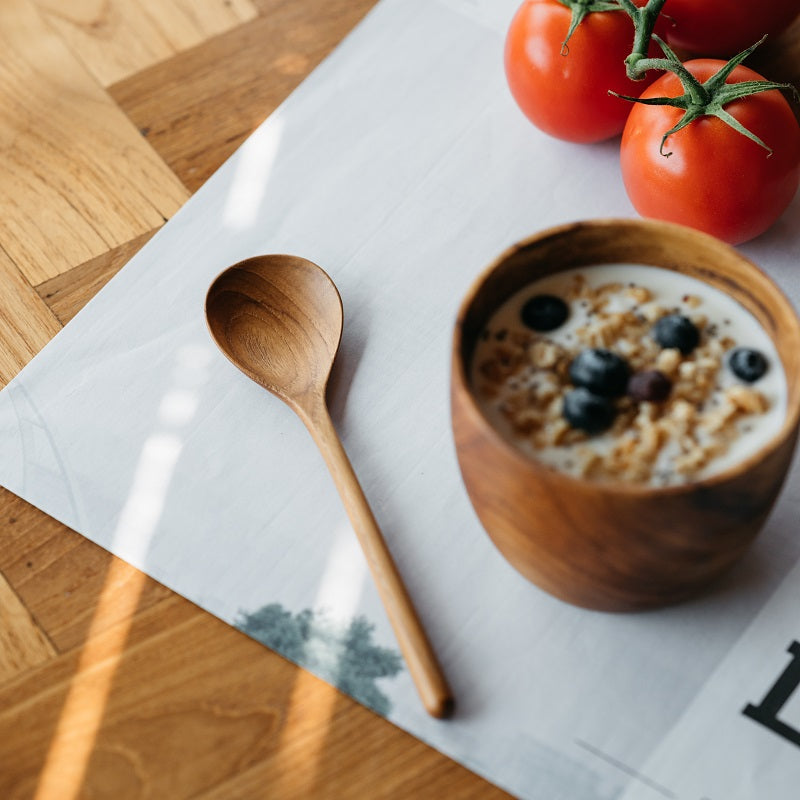 A person is holding a wooden spoon above a bowl of oatmeal topped with blueberries. The bowl is placed on a green plate on a wooden table.