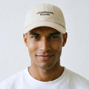 A man wearing a beige baseball cap with the text 'CHAMPAGNE, PLEASE.' embroidered in black on the front, paired with a white crewneck t-shirt against a plain light background.