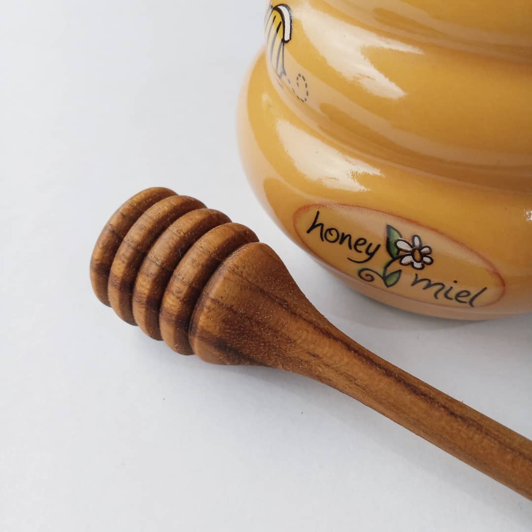 A handcrafted Teak Wood honey dipper lying on a white surface, next to a yellow honey jar with a floral pattern.