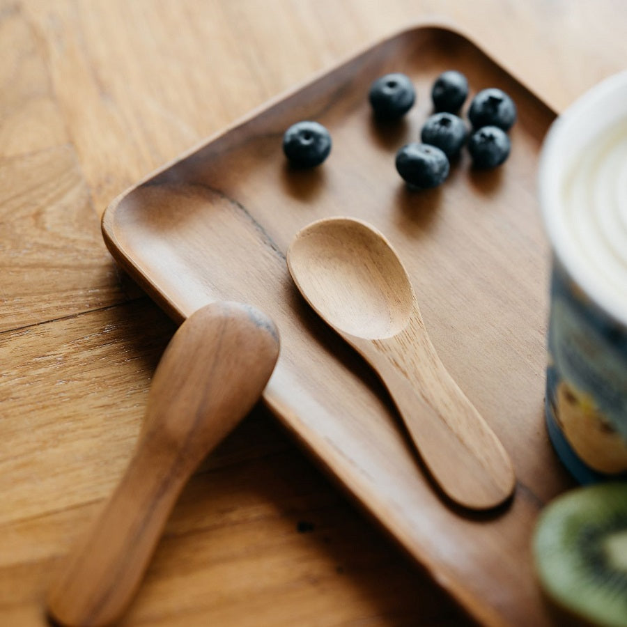 A person holding a teak wood ice cream spoon above a bowl of ice cream, with fruits and a wooden board in the background.