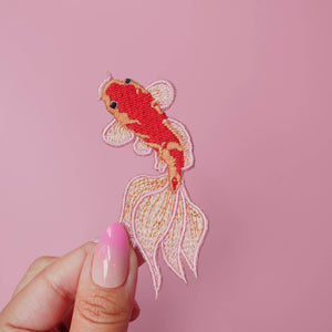 Embroidered goldfish patch held by a hand against a pink background