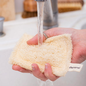 Person holding a loofah sponge under running water in a bathroom setting.