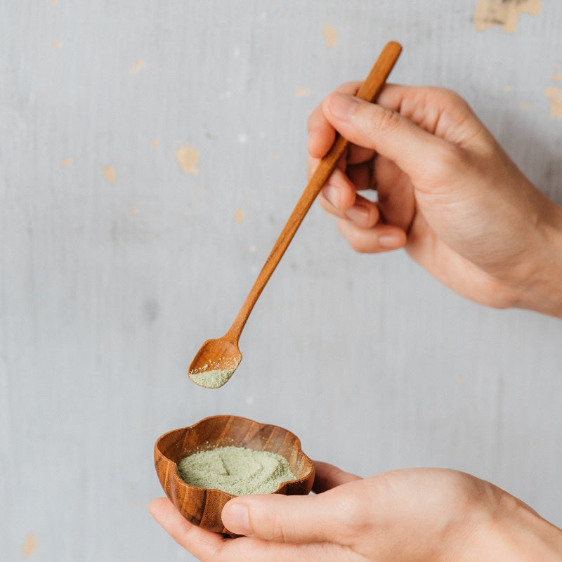 A person is holding a small wooden bowl filled with matcha powder, with a matcha spoon about to scoop some powder.