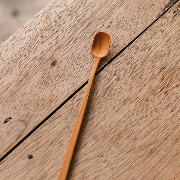A person is holding a small wooden bowl filled with matcha powder, with a matcha spoon about to scoop some powder.