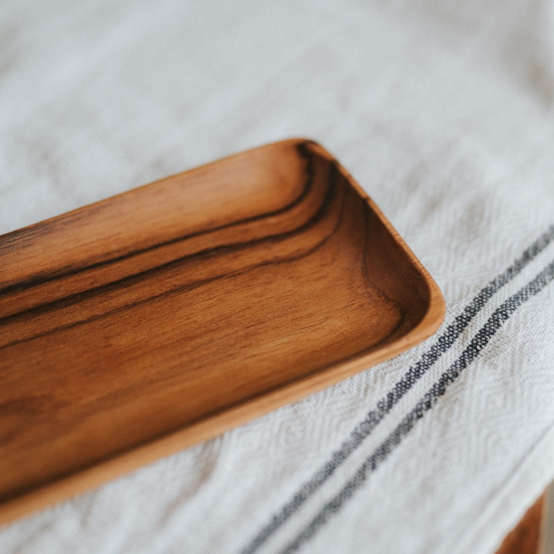 A rectangular teak wood plate placed on a table with a blue textured cloth, displaying various food items like rolls and sweets.