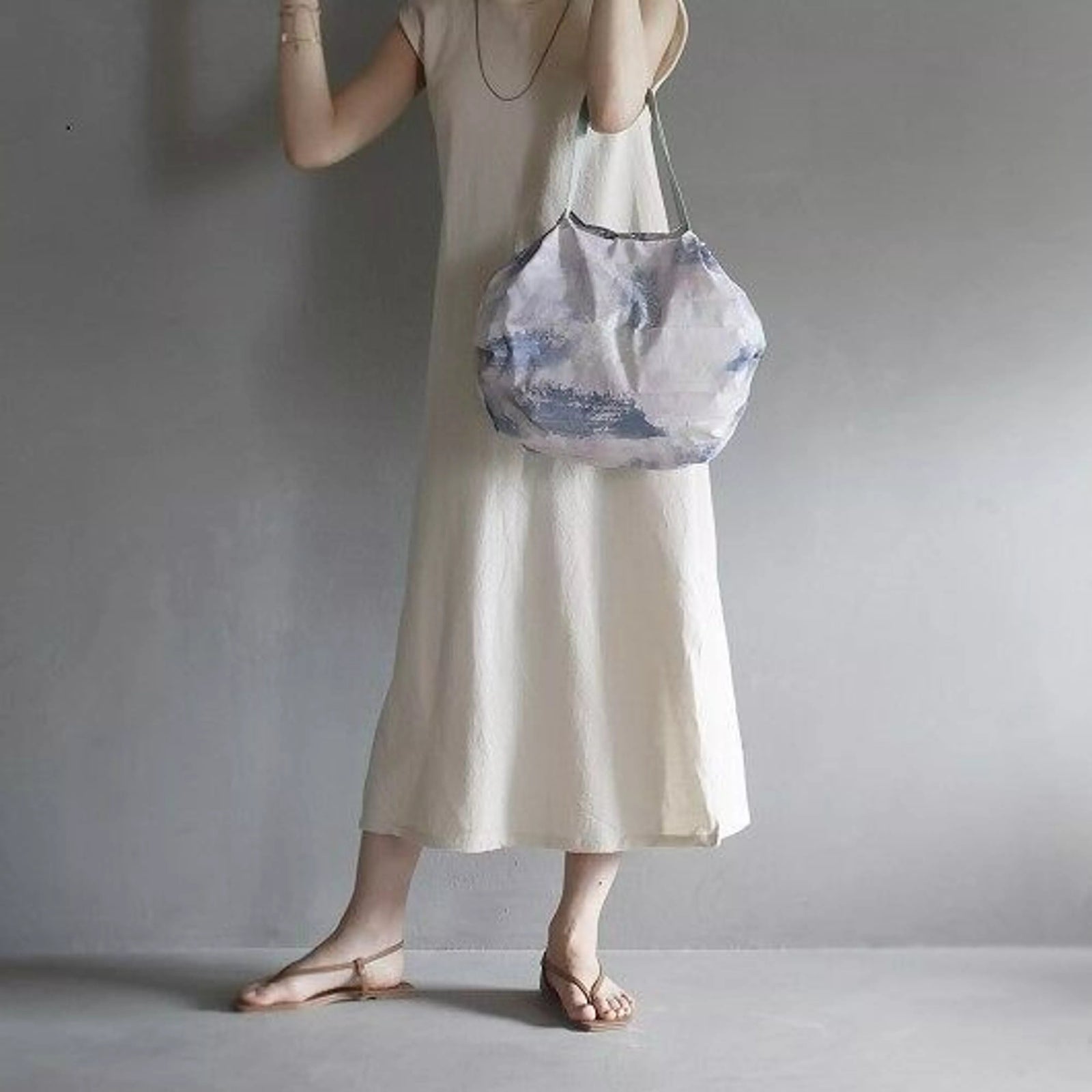 Person wearing a long light-colored dress holding a patterned bag against a plain background