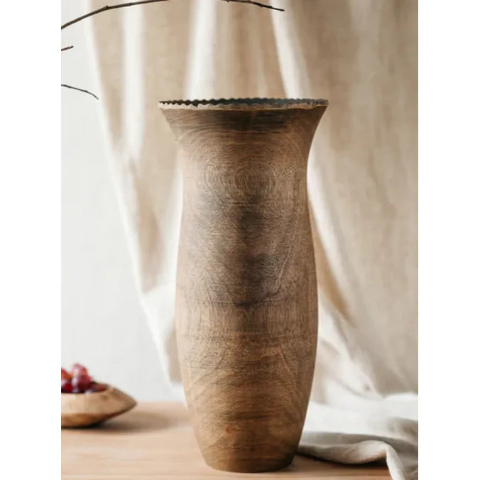 A tall, rustic mango wood vase with a flared top, displayed against a soft, draped fabric background.