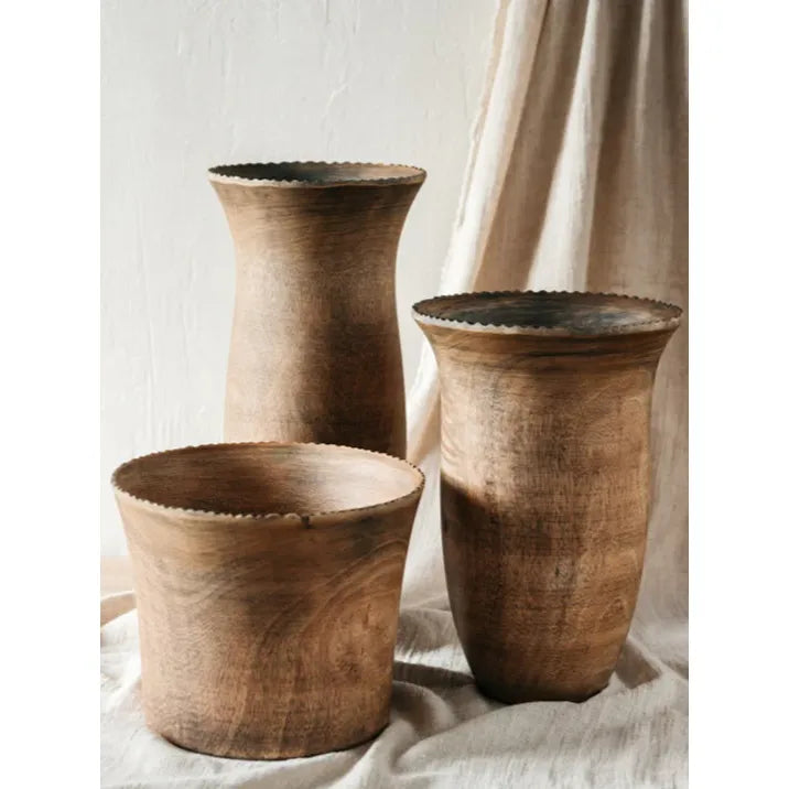 A tall, rustic mango wood vase with a flared top, displayed against a soft, draped fabric background.