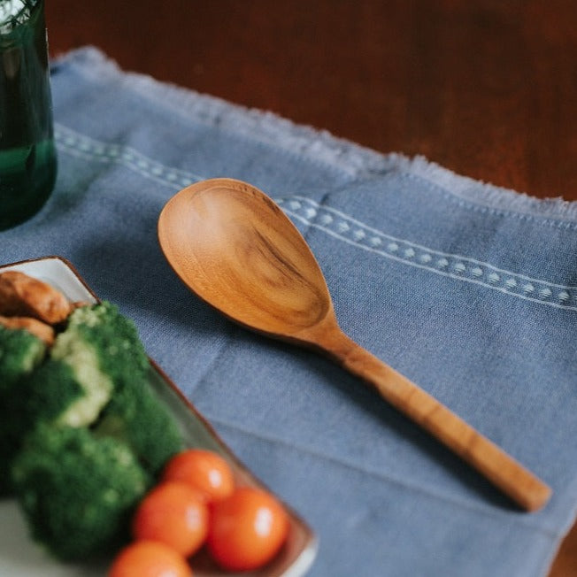 A handcrafted solid Teak Wood serving spoon with a smooth finish, placed on a blue textured surface, with vegetables and a glass bottle in the background.