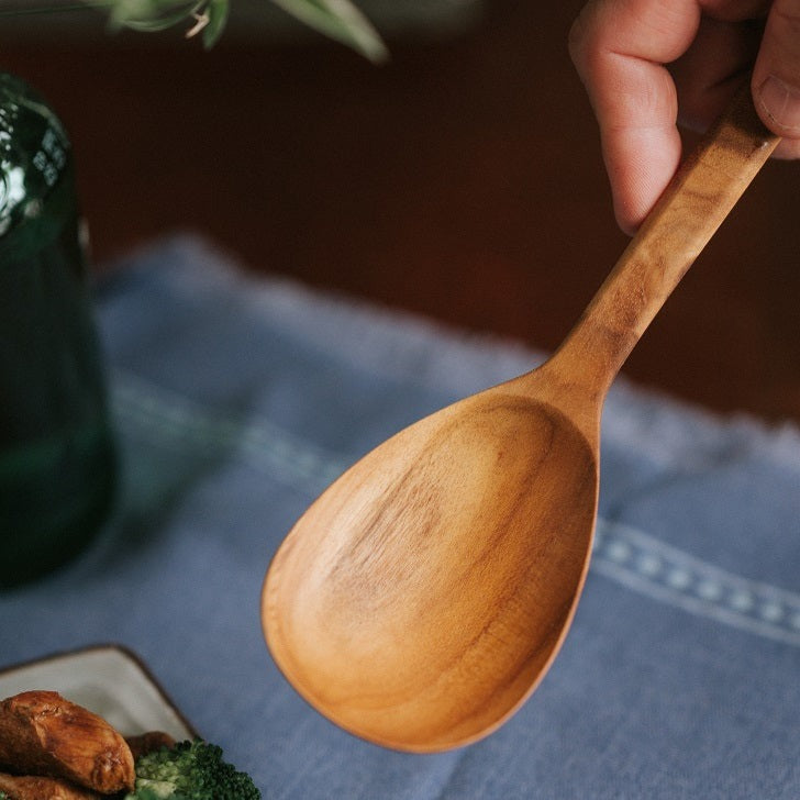 A handcrafted solid Teak Wood serving spoon with a smooth finish, placed on a blue textured surface, with vegetables and a glass bottle in the background.