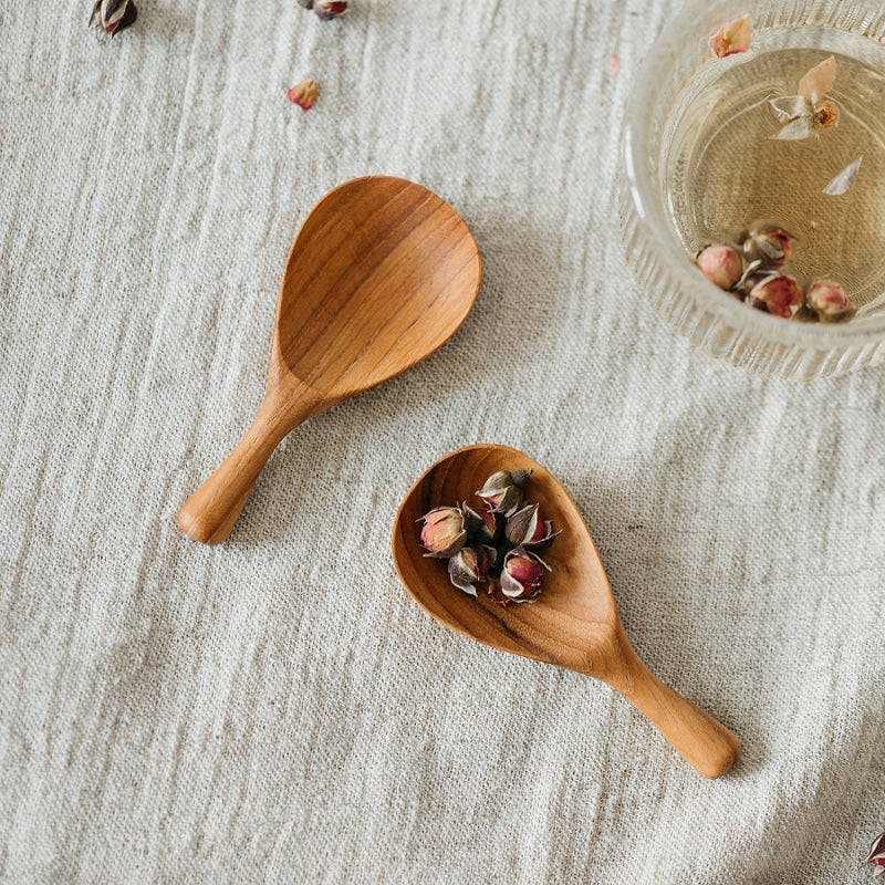 Two wooden spoons on a textured fabric surface, one spoon is empty and the other is holding small dried flowers.