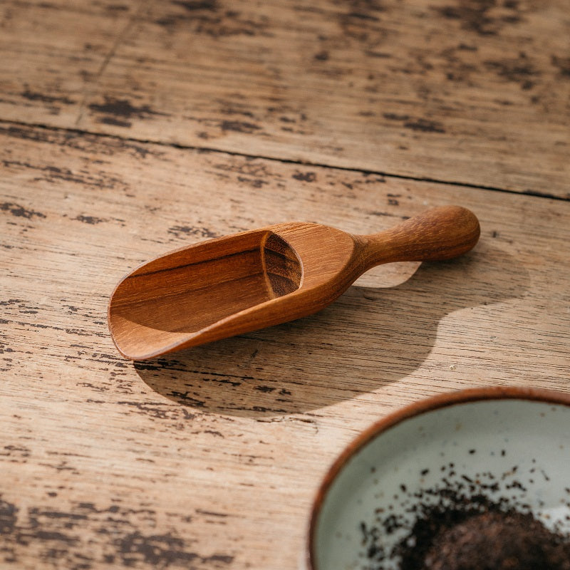 A handcrafted solid Teak Wood tea leaf scoop placed on a wooden surface, with a bowl of tea leaves visible in the background.