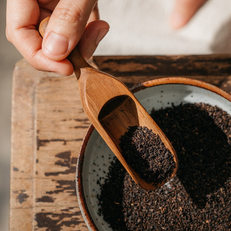 A handcrafted solid Teak Wood tea leaf scoop placed on a wooden surface, with a bowl of tea leaves visible in the background.