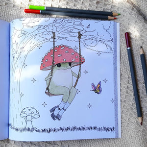 An open coloring book or sketchbook lying on a textured beige fabric surface, showing a whimsical line-art illustration of a frog sitting on a mushroom swing under a red spotted mushroom cap. Several colored pencils are placed around the book.