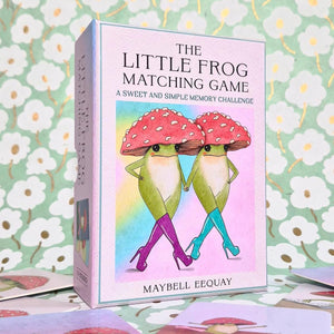 The Little Frog Matching Game box standing upright on a surface with matching cards scattered around it. The box features an illustration of two cartoon frogs wearing red mushroom hats and colorful high heels against a pastel rainbow background.