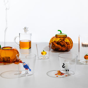 A set of Halloween-themed clear glass tableware on a white surface, including a pumpkin-shaped candle holder, a ghost-topped mug with amber tea, a small pumpkin jar, and several glasses with animal and pumpkin illustrations.