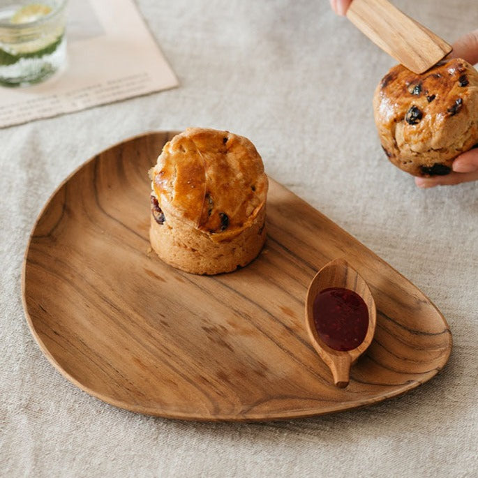 Two teak wood plates on a grey fabric surface, one plate has a muffin topped with a brown sugar and cranberry mixture, and the other has a dollop of red jam. A person is in the background, partially visible, cutting a piece of the muffin with a fork.