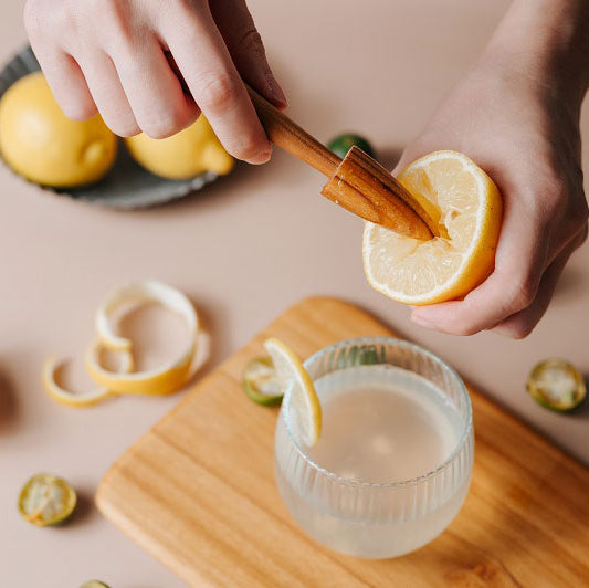 A person's hands using a teak wood lemon squeezer to juice a lemon, with the squeeze of lemon juice into a glass, surrounded by sliced lemons and a wooden board on a kitchen counter.