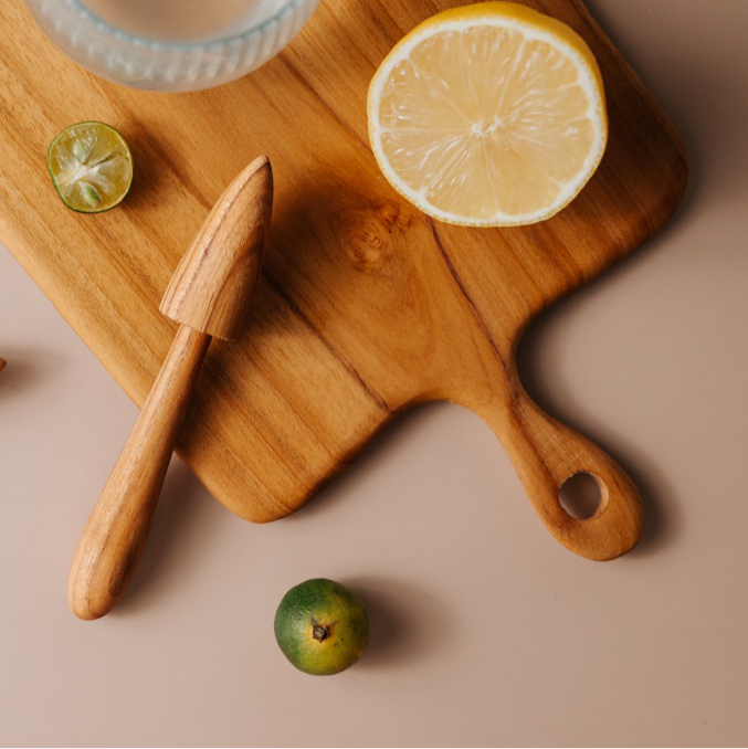 A person's hands using a teak wood lemon squeezer to juice a lemon, with the squeeze of lemon juice into a glass, surrounded by sliced lemons and a wooden board on a kitchen counter.