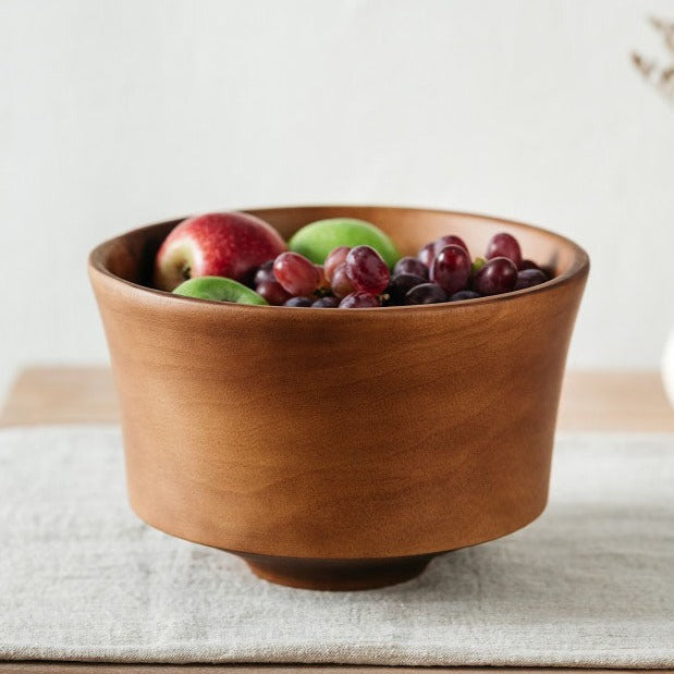 A wide, hand-turned mango wood fruit bowl on a linen mat, filled with various dried fruits.