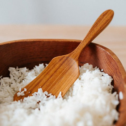A Teak Wood rice/potato salad scoop resting in a bowl filled with rice. The scoop has a golden brown color with a smooth finish and a handle for easy grip.
