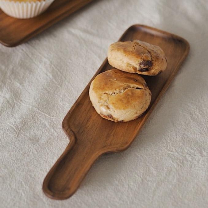 A teak wood serving board with a handle, with two muffins or scones placed on it, set against a linen background.